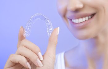 A person smiling holds a clear dental aligner against a light purple background.